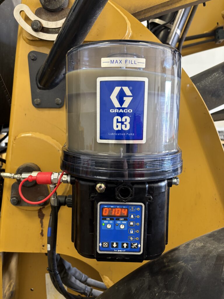 Close-up of a Graco G3 lubrication pump mounted on yellow machinery, installed by industrial equipment services Sault Ste. Marie. The transparent reservoir is filled with lubricant, and the digital control panel displays 109, with various wires connected.