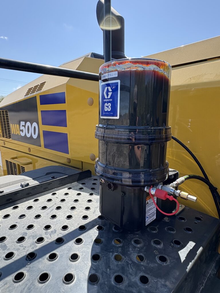A close-up of a Graco G3 grease pump mounted on the perforated metal platform of a yellow Komatsu WA500 heavy equipment vehicle, with blue sky visible in the background.