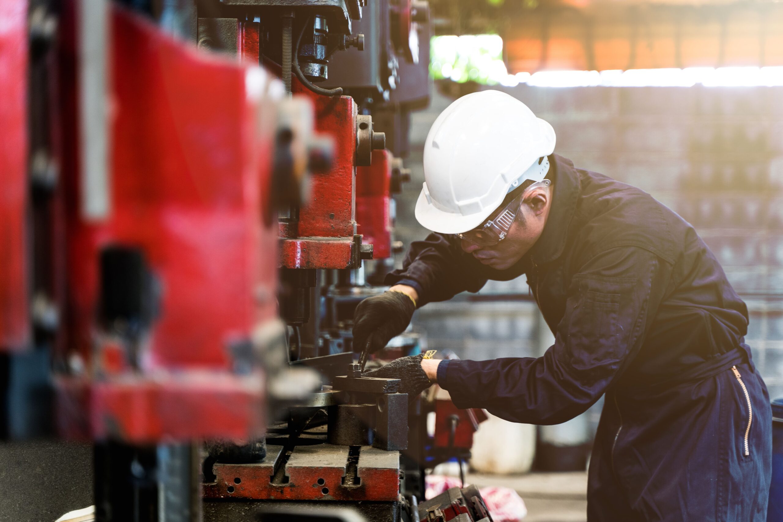 A person wearing a white safety helmet, safety glasses, and dark coveralls operates machinery in an industrial equipment service setting in Sault Ste. Mar, working with metal parts using gloved hands.