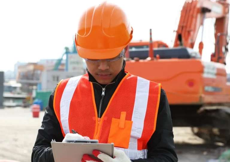 A construction worker in an orange safety helmet and vest writes on a clipboard at a Sault Ste. Marie worksite with heavy machinery and fire suppression systems in the background.