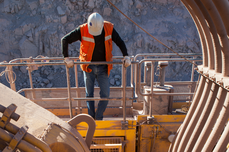 A worker wearing a white hard hat, orange safety vest, and gloves stands on a metal platform at a rocky industrial site in Sault Ste. Marie, holding a railing and inspecting the area for fire suppression systems below.