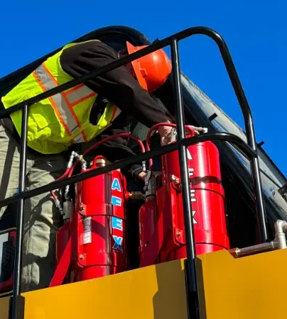A worker in a safety vest and orange helmet inspects red fire suppression systems on industrial machinery in Sault Ste. Marie, framed by a clear blue sky.