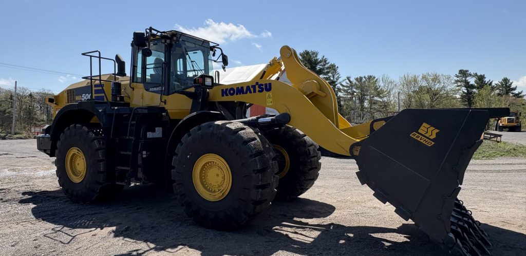 A large yellow and black Komatsu front loader with a metal bucket attachment is parked on a gravel lot under a clear blue sky, surrounded by some trees and construction equipment in the background.