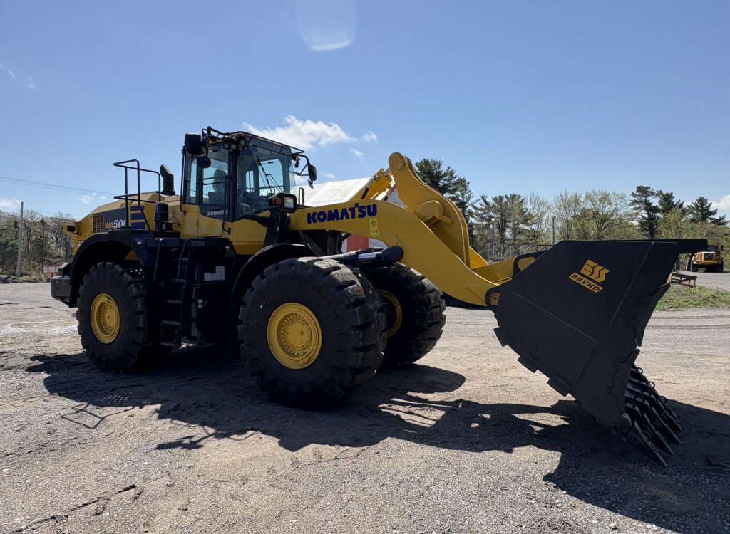 A large yellow and black Komatsu front loader with a metal bucket attachment is parked on a gravel lot under a clear blue sky, surrounded by some trees and construction equipment in the background.