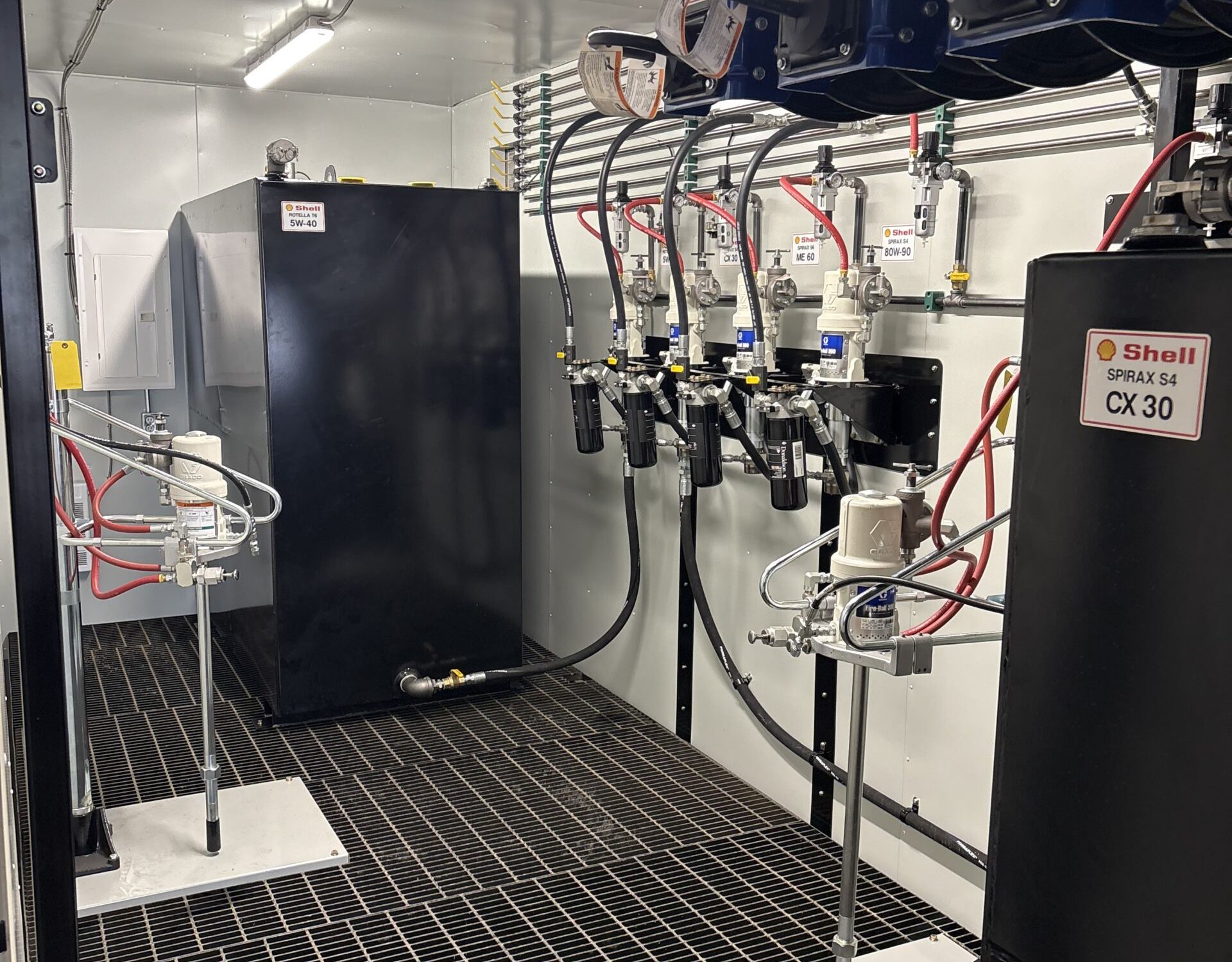Interior view of an industrial lubrication room in Sault Ste. Marie featuring black storage tanks, specialty equipment components, Shell-branded oil dispensers, metal pipes, gridded flooring, labeled hoses, and visible electrical wiring on the wall.