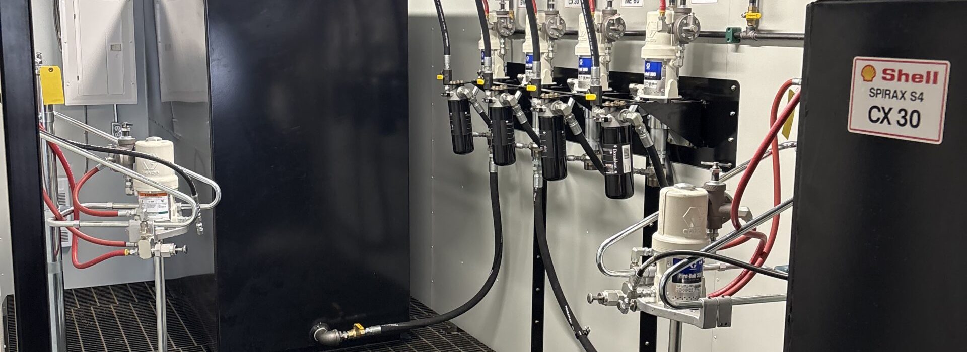 Interior of an industrial lubricant storage room with black tanks, dispensing equipment, hoses, and Shell oil containers labeled CX 30 and 80W-90, all arranged on a grated metal floor.