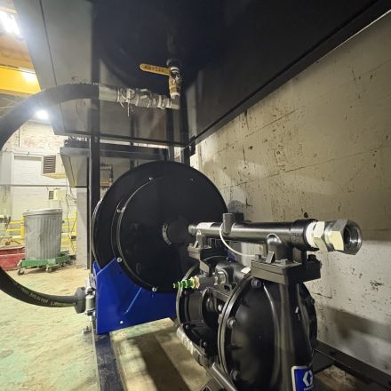 A close-up view of industrial equipment, including a black hose reel, metal pipes, and a pump on a blue base, set against a wall in a workshop or factory with worn flooring.