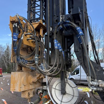 A construction worker in safety gear operates underneath a large drilling machine with hydraulic hoses, set up on a construction site with trees and a white van in the background.
