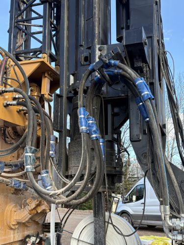 Close-up of heavy construction machinery with several thick, black hydraulic hoses wrapped in blue tape, connected to large metal parts. A white van and trees are visible in the background.