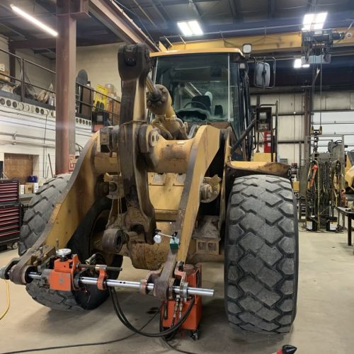A large yellow front-end loader is parked inside a workshop. Its front wheels are removed and alignment equipment is attached to the hubs. Tools and machinery are visible around the workspace.