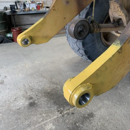 Two yellow metal arms, likely industrial service equipment in Sault Ste. Marie, are shown with new metal bushings installed. A large tire and various workshop tools and containers are visible in the background.
