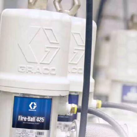 Close-up of two Graco Fire-Ball 425 oil pumps, a staple in industrial service equipment Sault Ste. Marie, showing their white exteriors, company logo, blue labels, and connected hoses and pipes in a workshop setting.